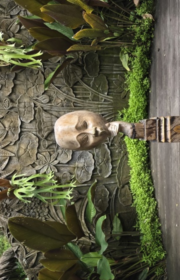 A carved wooden sculpture of a face stands on a decorative pedestal, framed by lush tropical plants and a stone floral relief wall.