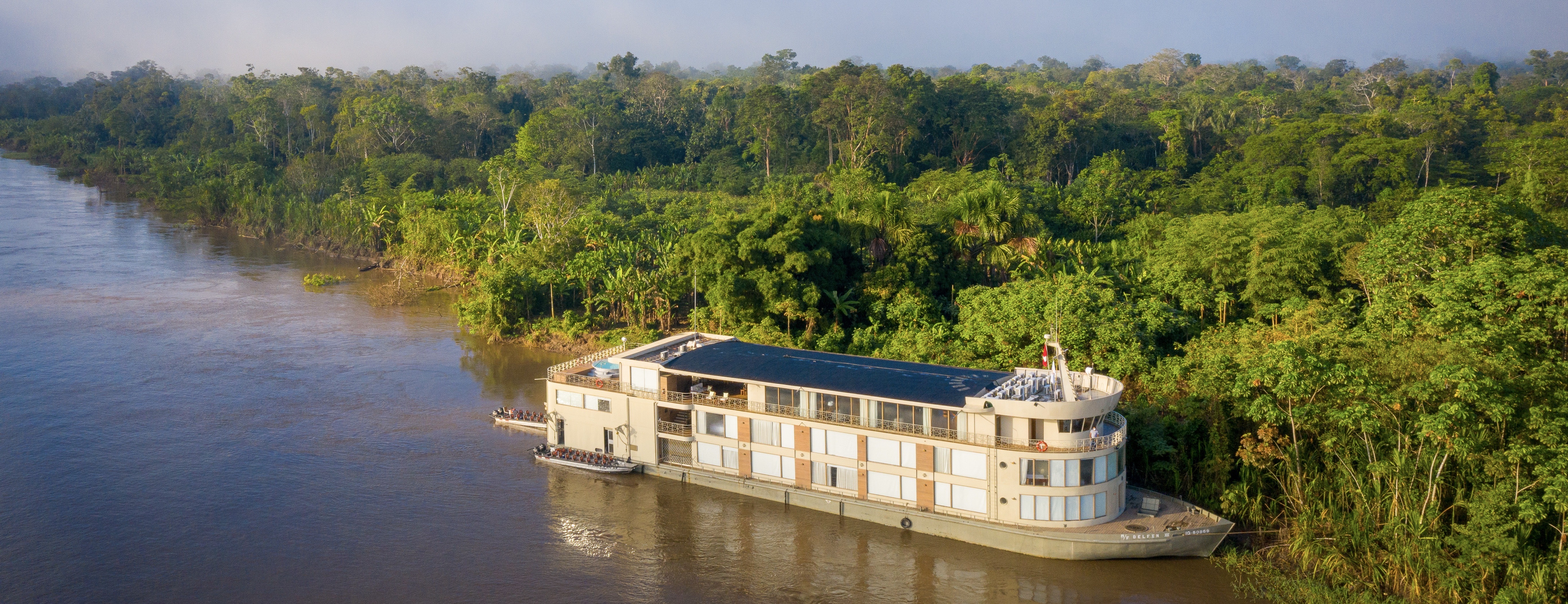 Aerial view of the Delfin III luxury river cruise gliding through the lush green Amazon rainforest in Peru. 