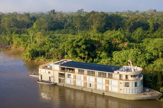 Aerial view of the Delfin III luxury river cruise gliding through the lush green Amazon rainforest in Peru. 
