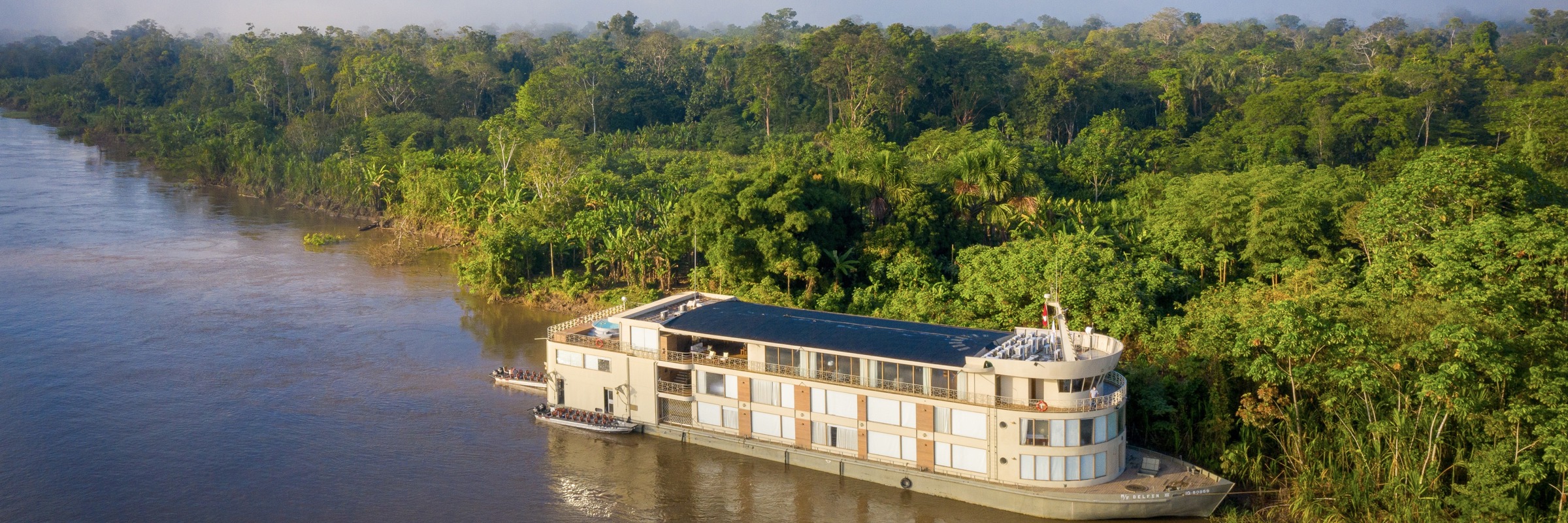 Aerial view of the Delfin III luxury river cruise gliding through the lush green Amazon rainforest in Peru. 